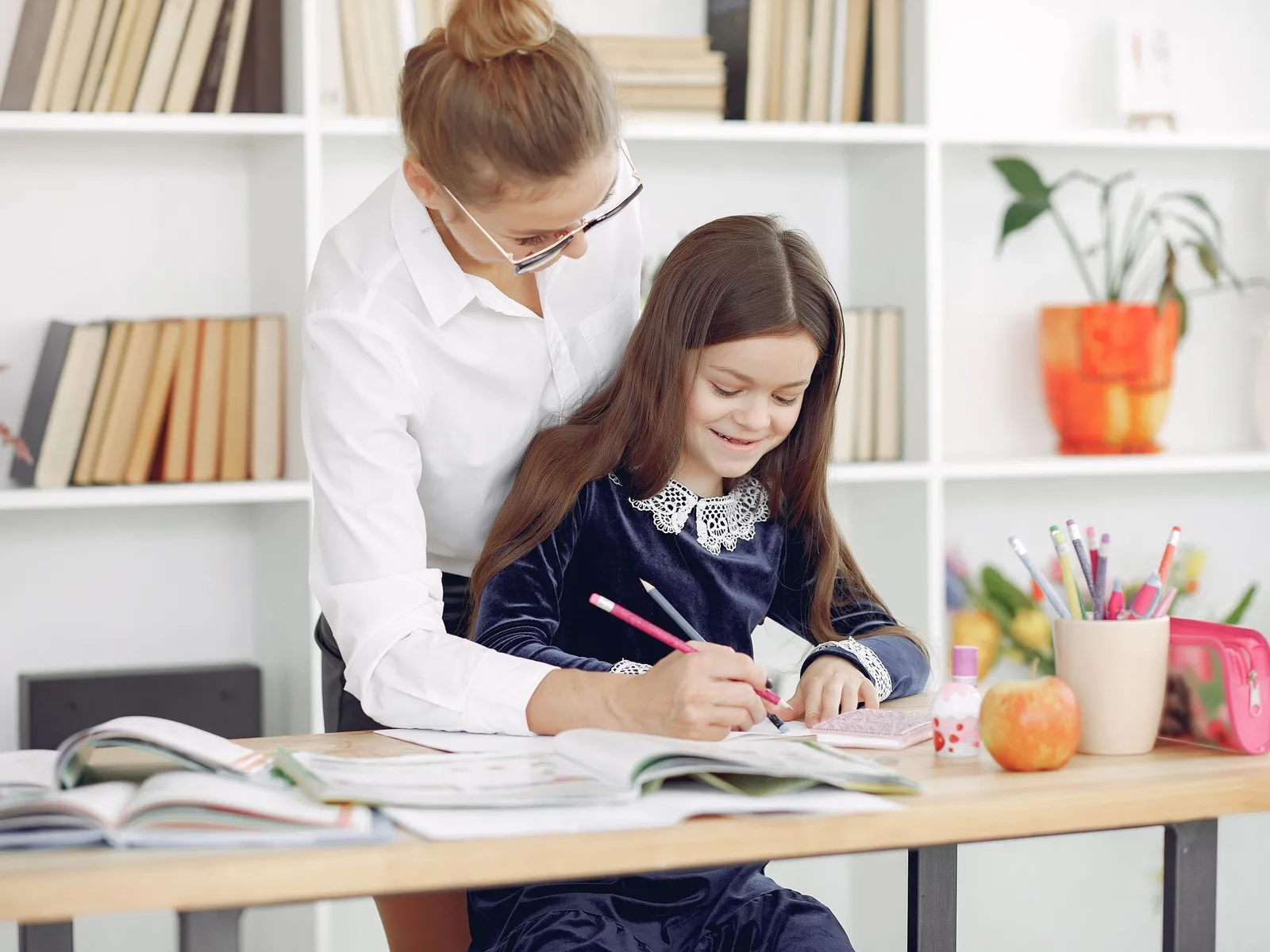 An adult helping a child write in a notebook at a classroom table.